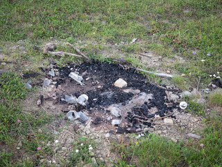 Abandoned campfire with ashes, charcoal, burnt wood and plastic waste on grassy ground, showing pollution and environmental damage in nature.