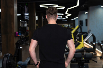 Naklejka premium Man standing in modern fitness gym. A man in a black t-shirt stands in a modern gym with exercise equipment in the background.