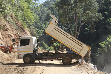 White dump truck diligently works, raised bed releasing earth onto rugged dirt road. Scene captures...