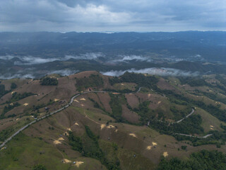 Naklejka premium Expansive aerial view captures verdant rolling hills beneath an overcast, dramatic sky. Patches of lighter earth mark areas of deforestation across green and brown terrain, showcasing an evolving