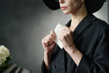 Middle aged Caucasian woman holding tissue with both hands standing near coffin, wearing dark...