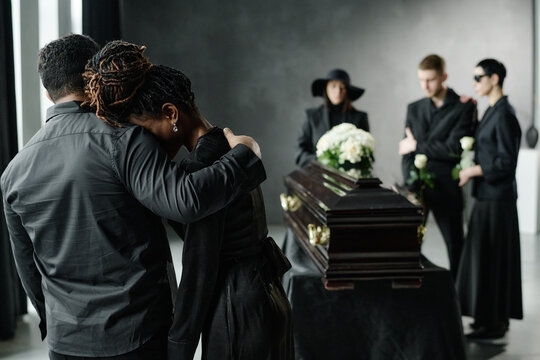 Black woman leaning on Black man for support while grieving near coffin, group of young adults and middle aged woman standing in background holding flowers - Powered by Adobe
