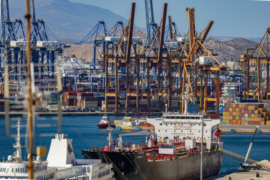 Aerial view of colossal cranes standing guard over cargo ships nestled in the bustling port, a symphony of industry against the azure backdrop, Piraeus, Greece.