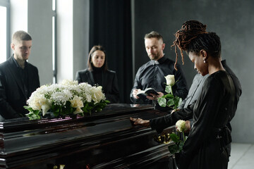 Group of middle aged and young adult men and women of diverse ethnicities standing around coffin holding roses, mourning and paying respects during funeral ceremony