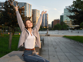 A cheerful woman enjoys a moment outdoors in a modern urban park, raising her arms with a wide smile, radiating confidence, happiness, and energy in a sunny, breezy setting.