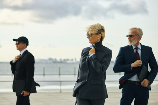 Caucasian middle aged woman in suit walking outdoors with two men in business attire and sunglasses, all holding briefcases and looking in different directions near waterfront