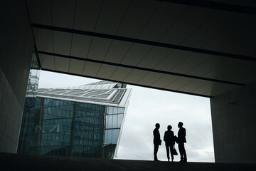 Three middle aged business professionals standing in modern urban setting, silhouettes engaged in conversation near glass office building, diverse group interacting outdoors © DragonImages