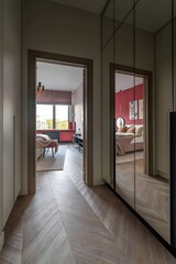 A modern hallway with mirrored wardrobe and wooden flooring leads to a bright living room with red accents.