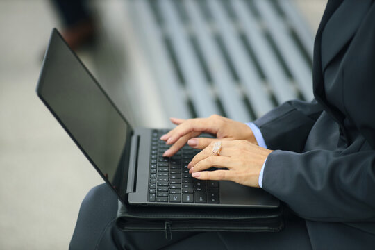 Middle aged businesswoman in formal suit typing on laptop outdoors, hands resting on keyboard, wearing business attire, sitting on bench, focusing on work, professional setting - Powered by Adobe