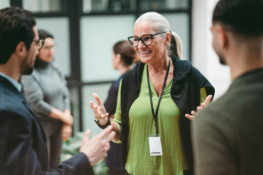 Smiling senior female business expert wearing eyeglasses discussing with colleagues at networking event in convention ce