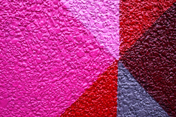 Multicolored textured wall with triangular patterns in vibrant shades of pink, purple, red, and gray. The surface has a rough, uneven aggregate texture with glossy finish.