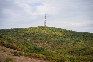 Obraz premium A red-and-white telecommunications tower rises above a forested hill, blending autumn foliage with modern infrastructure beneath a partly cloudy sky.