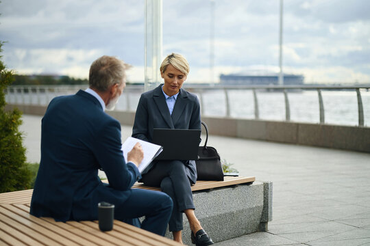 Middle aged Caucasian woman sitting outdoors using laptop while male colleague is reviewing documents on clipboard, both dressed in business attire near waterfront promenade