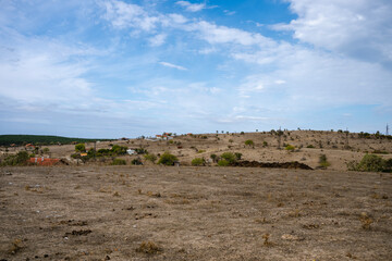 Scattered houses rest on a dry hillside with sparse trees and grass, under a partly cloudy sky in a semi-arid rural landscape.