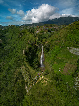 Aerial view of the Surodipo Waterfall cascading down a lush green valley, framed by rolling hills and a vibrant blue sky, Temanggung, Central Java, Indonesia.