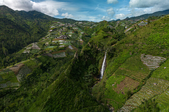 Aerial view of the cascading Surodipo Waterfall cutting a path through a vibrant green, terraced landscape under a cloudy sky, Temanggung, Central Java, Indonesia.