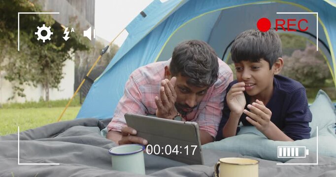 Father and son lying on blanket in backyard watching suspense clip on tablet tech, flinching
