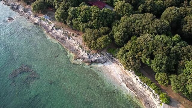 Aerial drone view of a rocky sunset coastline Lanterna peninsula in Istria, Croatia, with crystal clear turquoise water, surf waves, stone steps to the shore, and lush green forest above the cliffs.