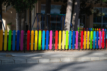 A vibrant rainbow-colored fence surrounds a playful outdoor playground, set beneath leafy trees and beside a city building.