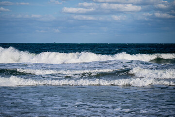 Waves crash and roll across the deep blue sea beneath a partly cloudy sky, capturing the raw energy and rhythm of the sea.