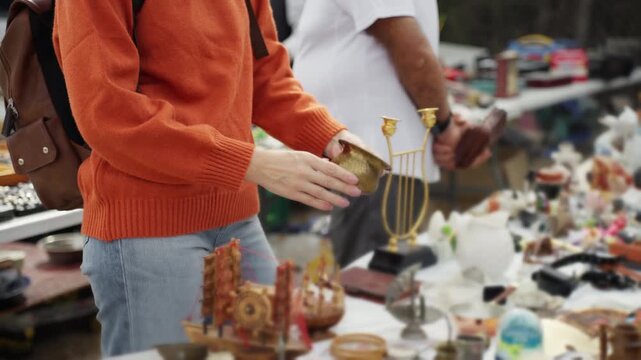 Woman browsing vintage trinkets at an outdoor flea market, inspecting items on stall table for bargains and antiques