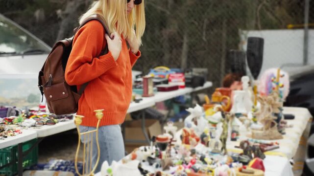 Backpack-wearing young woman browsing vintage and second-hand goods at an outdoor flea market searching for treasures. Europe travel. Lifestyle, vacation, tourism,nature, active life