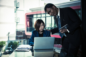 Asian businesswoman looking stressed while working on laptop as male colleague stands over her shoulder, speaking. The woman looks visibly uneasy and annoyed © Meng_Dakara