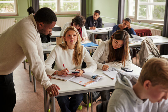 Male teacher leaning on desk near teenage female student solving math problem in classroom at school