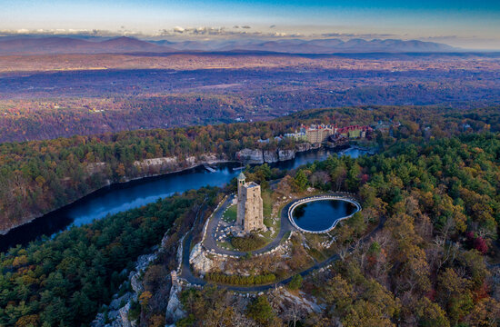 Aerial view of the stark stone tower of the Mohonk Preserve standing tall against the vibrant autumnal foliage and the tranquil waters of the lake, New York, United States.