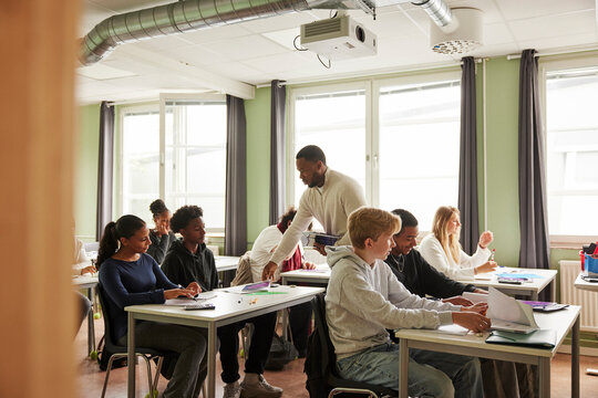 Male teacher distributing books to students in classroom at school