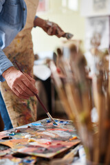 Oil painting techniques. Close-up view of male artist in stained apron dipping paintbrush into oil paint on wooden multicolored palette