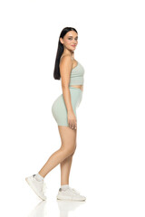 Studio shot of full length young woman in mint green activewear and white sneakers, walking in profile on white background.