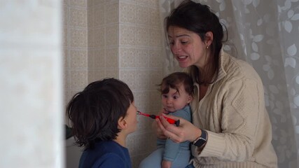 Mother helping young son brush teeth in bathroom while holding baby, multitasking parent caring for children during bedtime hygiene routine, family life at home - Powered by Adobe
