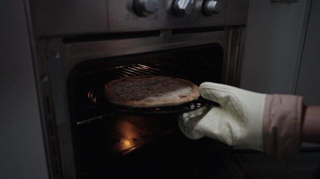 Close-up of male hand with oven mitt inserting frozen pepperoni pizza on perforated pan into preheated oven, preparing quick and easy meal for home cooking and dinner. Shooting in slow motion.