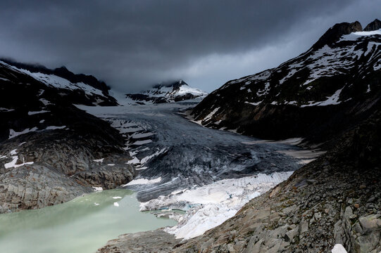 Aerial view of the Rh&Atilde;&acute;ne Glacier, a river of ice snaking through rugged terrain under a brooding sky, contrasted by the milky green glacial lake below, Obergoms, Valais, Switzerland.
