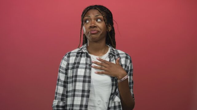 Woman with hand on chest, raised eyebrows and surprised expression wearing a plaid shirt in studio with pink backdrop; surprise curiosity.