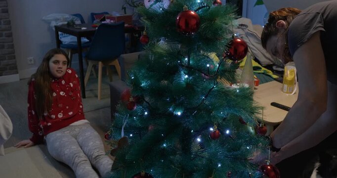 Daughter sitting on the floor, watching her mother decorate the Christmas tree. Tree in the foreground.