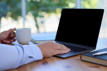 Man with cup of drink working on laptop at wooden table indoors, closeup