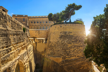 A huge limestone bastion with trees on top, historic stone walls and buildings under a bright blue...
