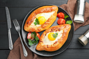 Tasty khachapuri with cheese and eggs served on black wooden table, flat lay