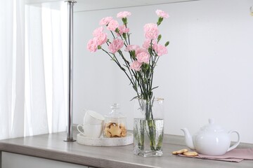 Beautiful carnation flowers in vase, cookies, teapot and cups on wooden cabinet indoors