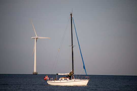 Sailboat with Danish flag navigates past an offshore wind turbine on calm seas.