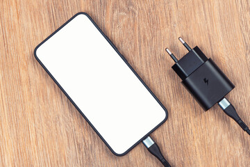Modern smartphone with blank white screen connected to black fast charger and USB-C cable, lying on wooden table. Concept of mobile technology, charging and digital devices