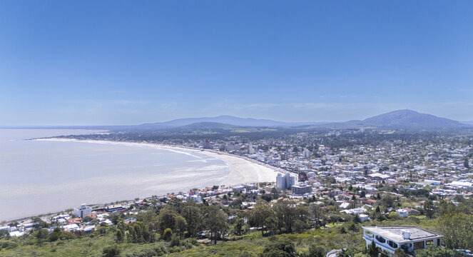 Aerial view of the sun-kissed beach curves gently along the coastline, contrasting with the vibrant city nestled against rolling hills, Piri&Atilde;&iexcl;polis, Maldonado Department, Uruguay.