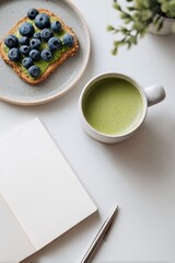 Flat lay of a ceramic plate with toast topped with mashed avocado and fresh blueberries next to a speckled mug of matcha latte, pen, notebook, and plant on a white desk