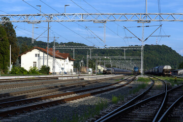 A freight train with a blue locomotive passes through a railway station in a hilly, green landscape under a sunny blue sky.