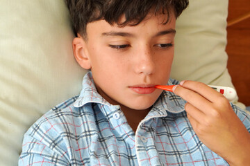 sick child in pajamas in bed, taking his temperature with a thermometer in his mouth