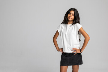 Strong teenage girl posing with hands on hips in casual outfit, white t-shirt and black denim skirt studio portrait.