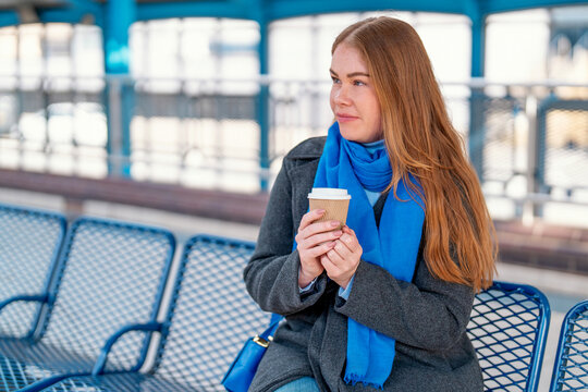 A woman stands at the train station on a cool morning, patiently waiting with a cup of coffee in hand - Powered by Adobe