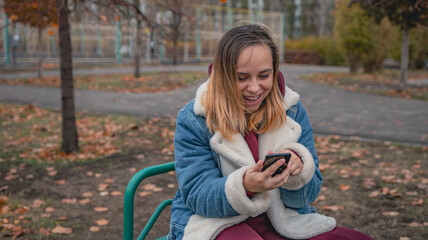 Obraz premium A woman dressed in a warm blue jacket sits on a bench in a city park, focused on her phone as autumn leaves fall around her on a cloudy day.